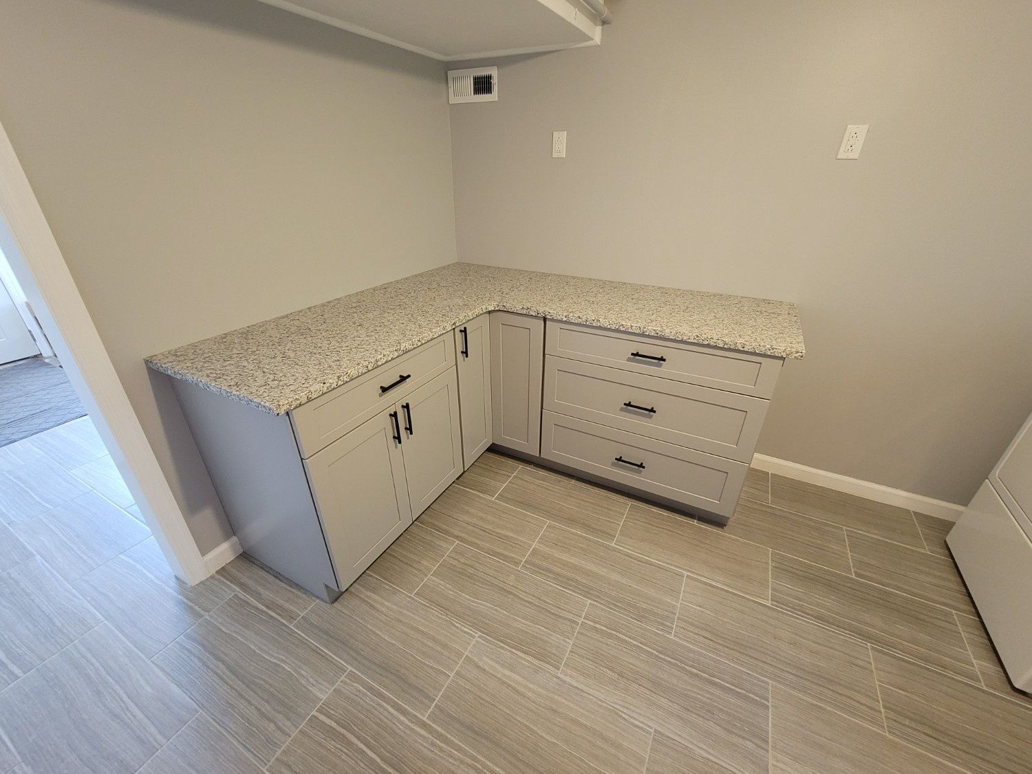 An L-shaped laundry room countertop with gray cabinets and drawers set against light gray walls and wood-look tile floors.