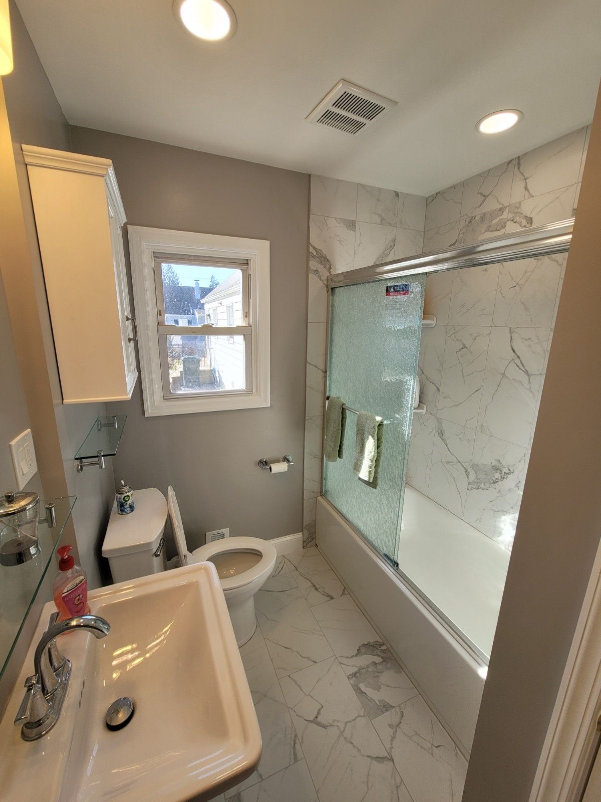 A bathroom featuring a gray wall, white marble-patterned shower tiles, a vanity sink, and a toilet under a window.