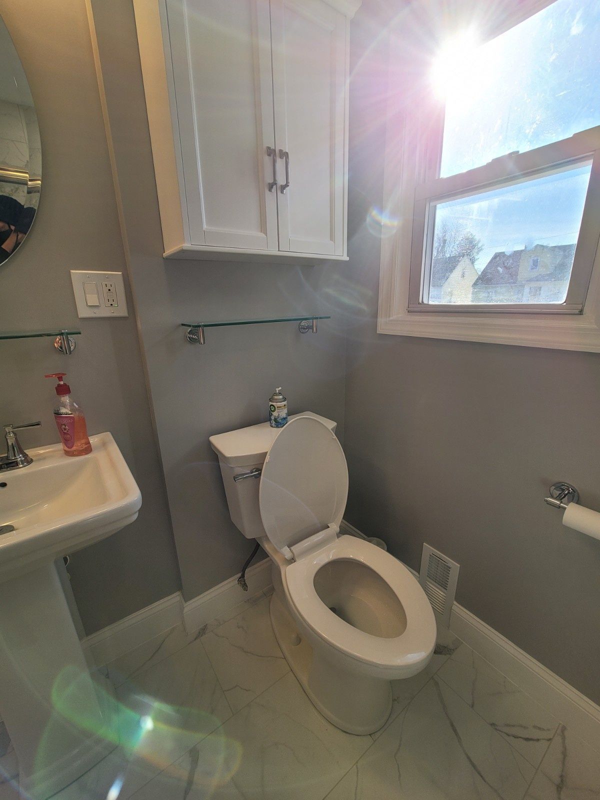 A small bathroom with a pedestal sink, white cabinet, toilet, and grey walls with natural light from a window.