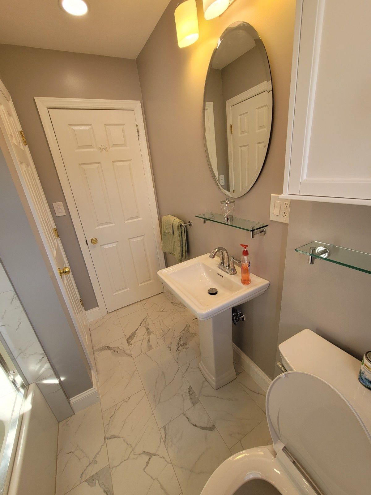 A modern bathroom with a pedestal sink, oval mirror, white door, and light-colored marble-patterned tile floor.