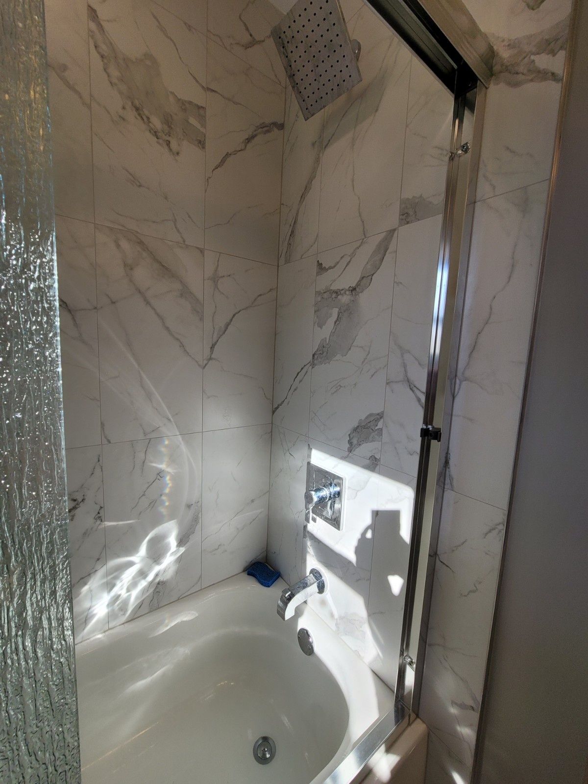 A marble-tiled shower alcove featuring a square rain showerhead, a chrome faucet, and a glass partition door.