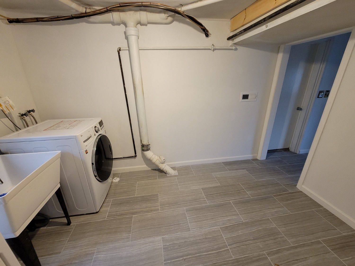 A laundry room with a washing machine, a utility sink, and white walls, featuring exposed plumbing and grey tiled floors.