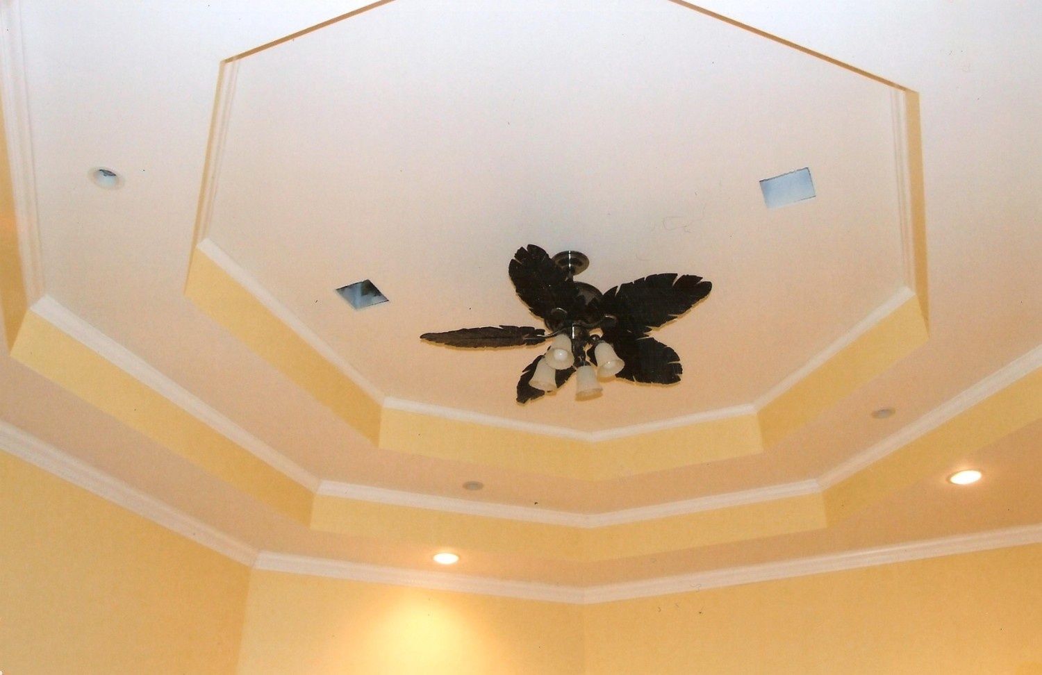 A low-angle view of a cream-colored octagonal tray ceiling with a dark brown ceiling fan and recessed lighting.