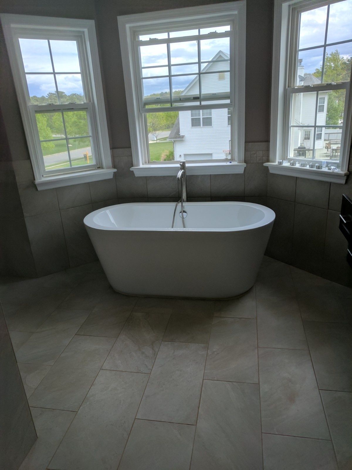 A white freestanding bathtub centered in front of three large windows in a room with light-colored tile floors and walls.