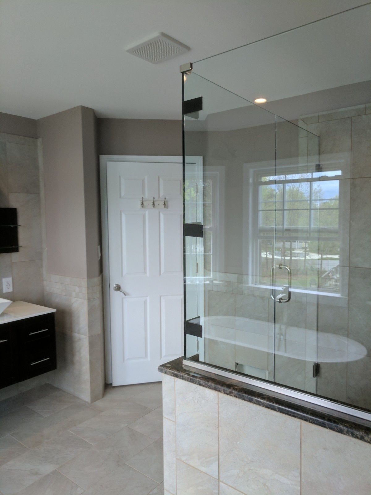 A modern bathroom featuring a walk-in glass shower, a white door, neutral tiled walls, and a dark wood vanity cabinet.