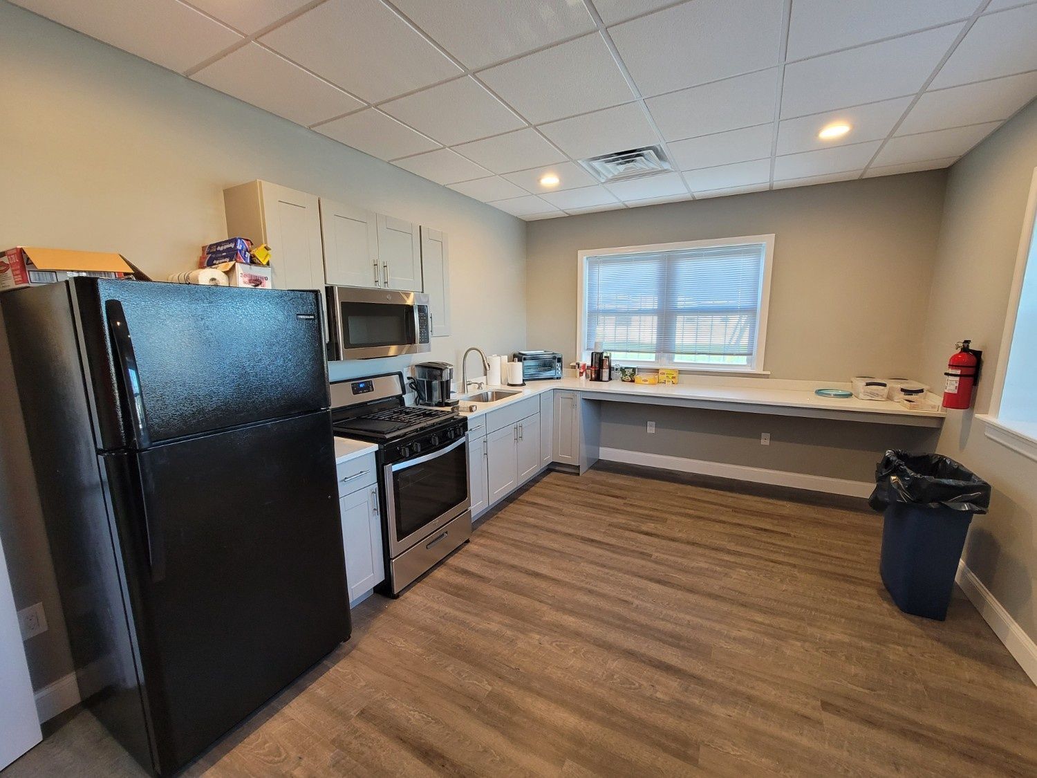 A kitchen area with a black refrigerator, a stove, a microwave, and a long counter along a wall with a window.