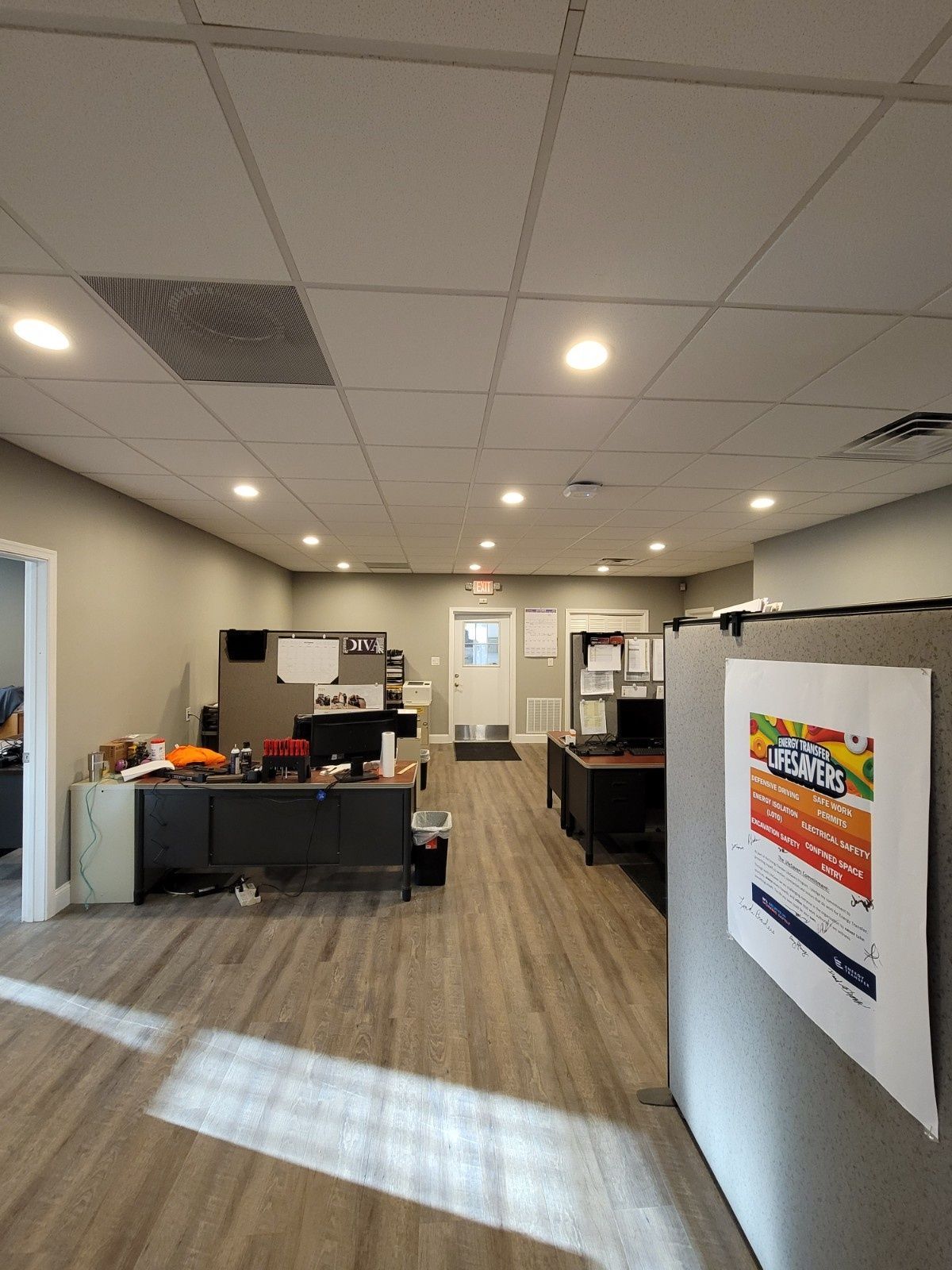 An office interior with wood-look flooring, multiple desks with cubicle partitions, a back door, and a sign on a partition.