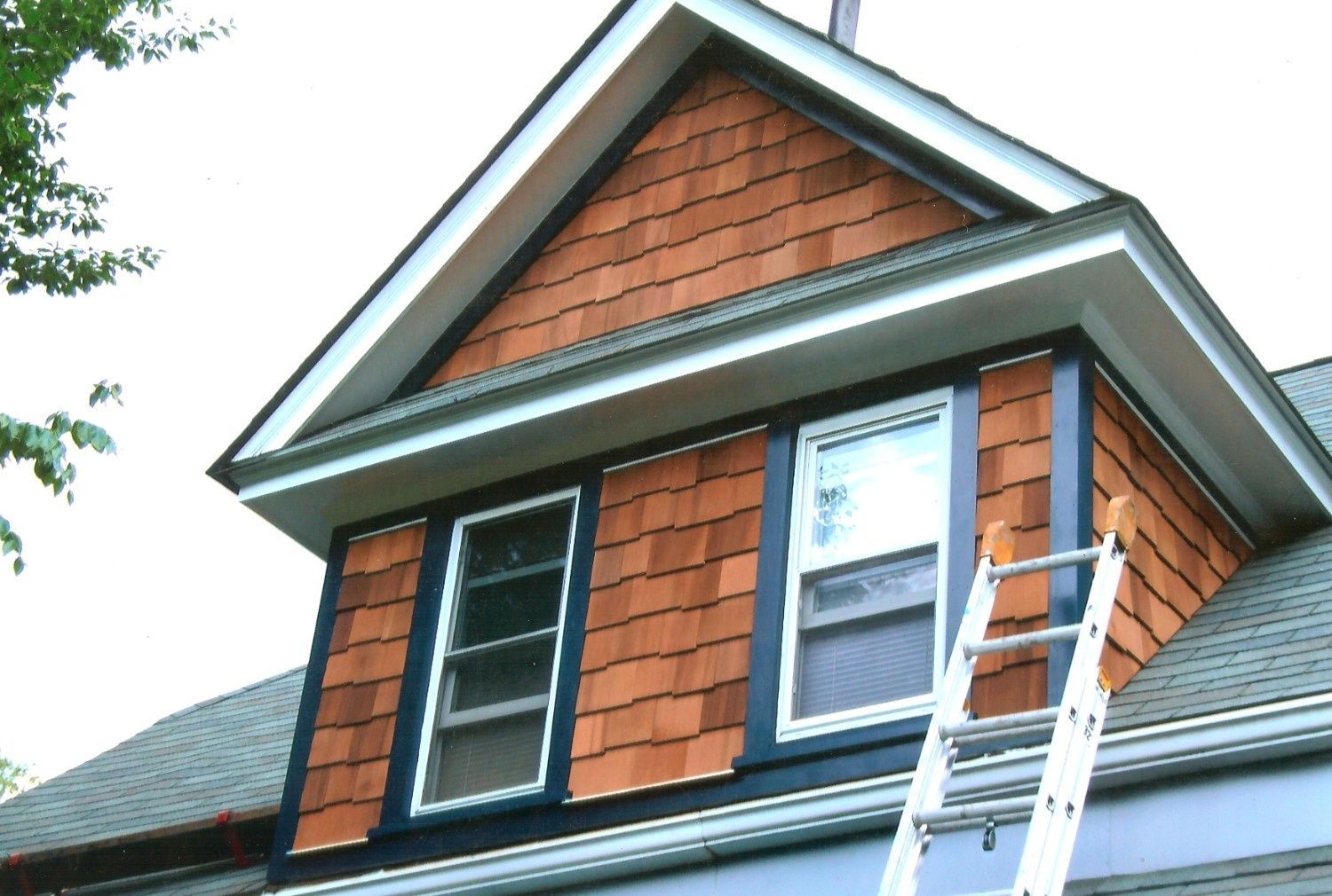 A close-up of a house dormer featuring cedar shingle siding and two windows, with a ladder leaning against the side.