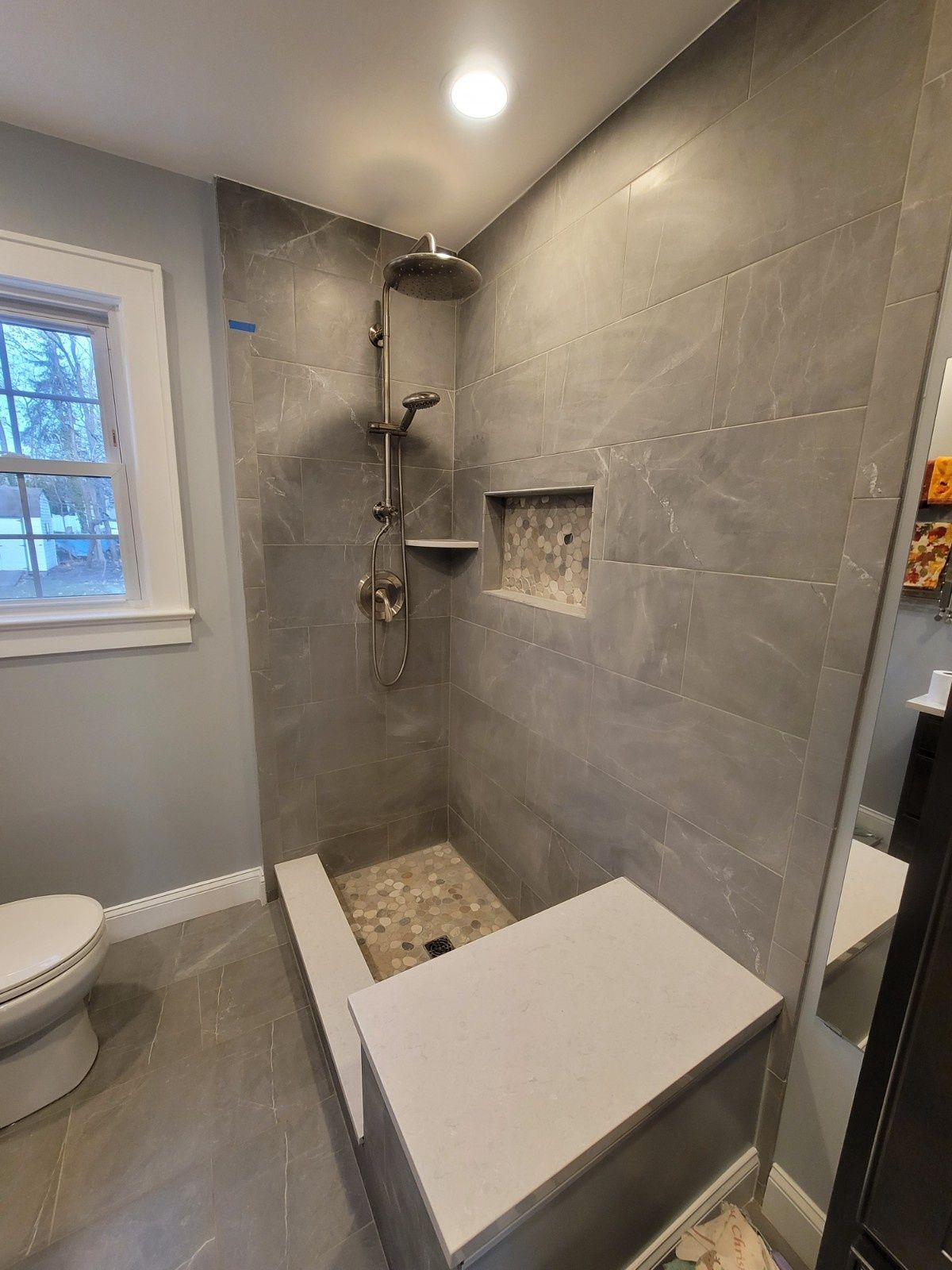 A walk-in shower with gray tiled walls, a bench seat, rainfall showerhead, and pebble floor tiling in a bathroom.