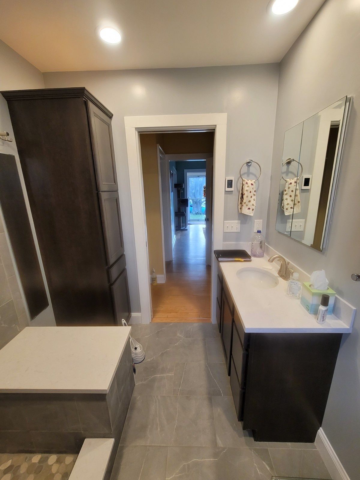 A modern bathroom featuring a dark vanity, a tall storage cabinet, grey tile floors, and a view into a hallway.