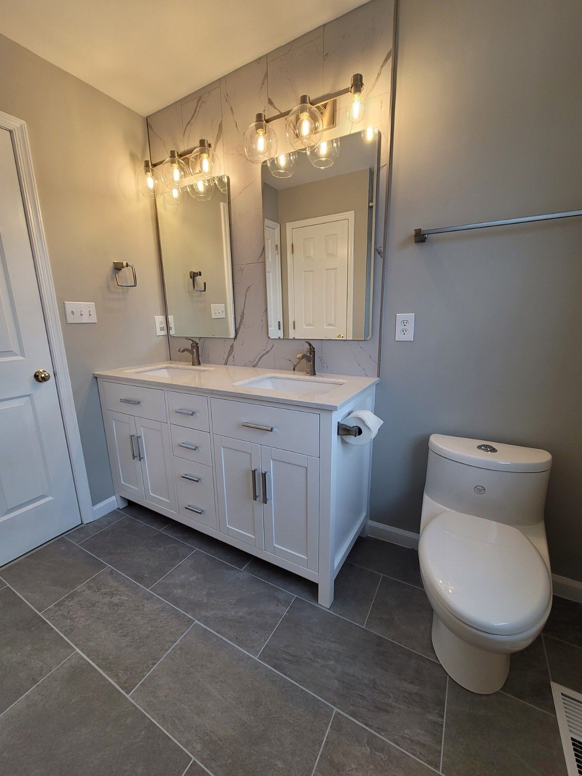 A bathroom vanity with white cabinets, dual sinks, and marble-look backsplash, illuminated by wall lights next to a toilet.