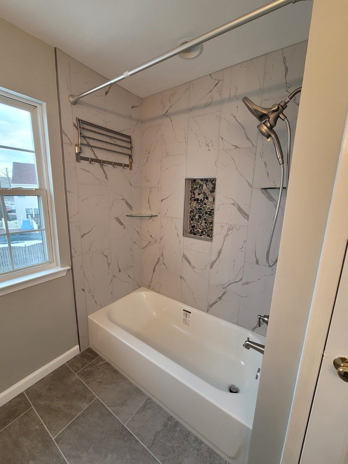 A bathroom featuring a white bathtub, marble-style tiled walls, a showerhead, a storage niche, and a window to the left.