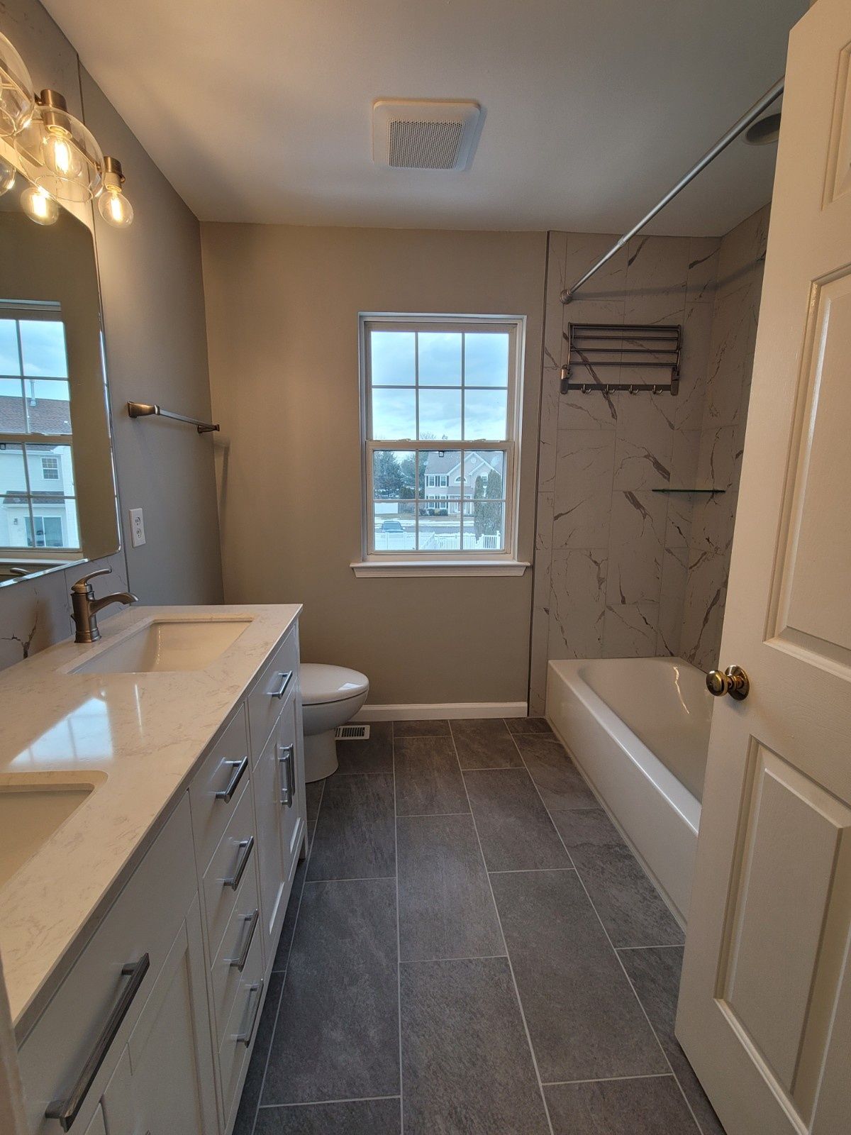 A modern bathroom with a double vanity, a window, a toilet, and a bathtub with marble-look wall tiles and grey floor tiles.