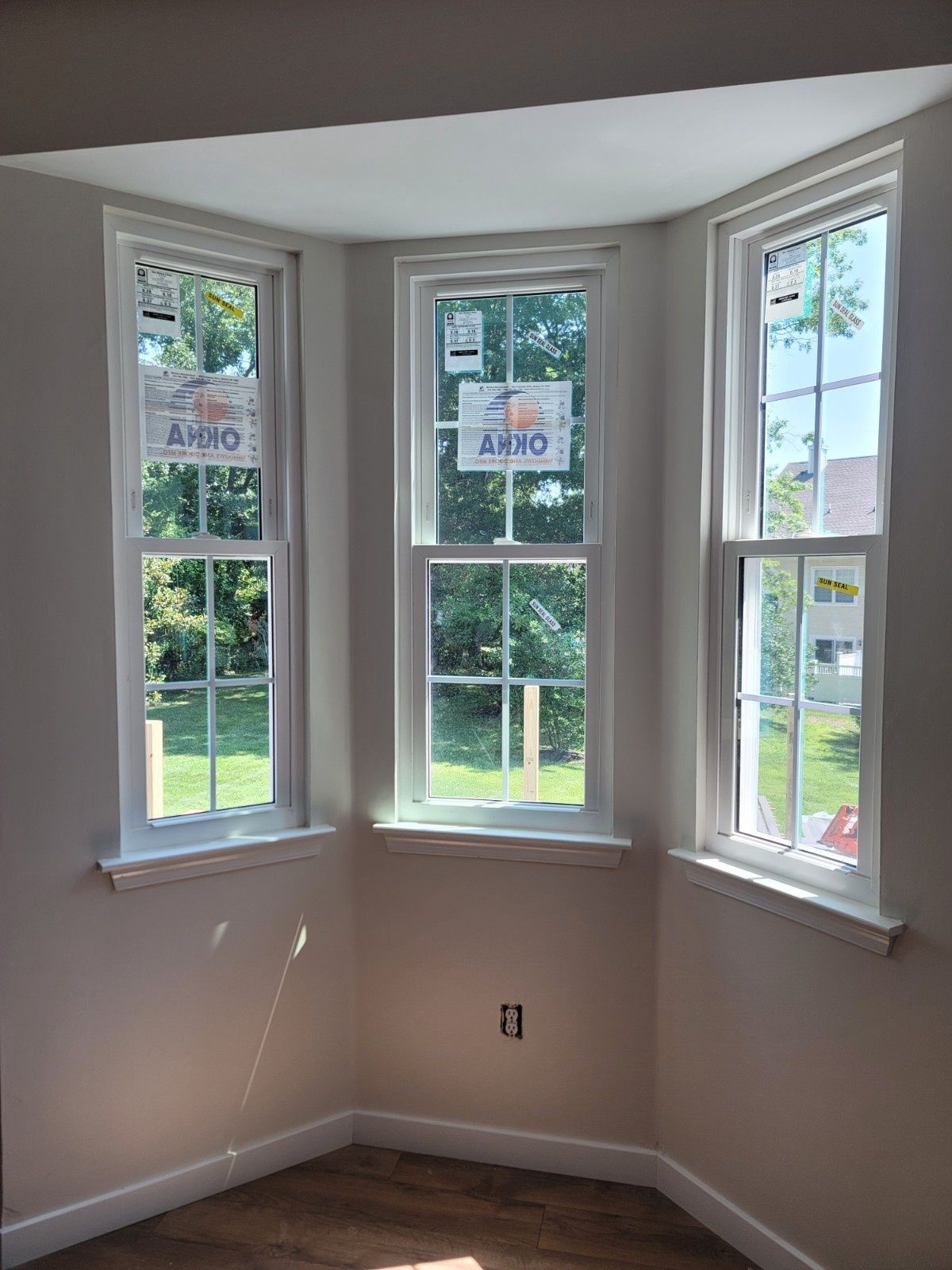 A three-paneled bay window with white trim, set in a light-colored room with wooden flooring and a visible wall outlet.