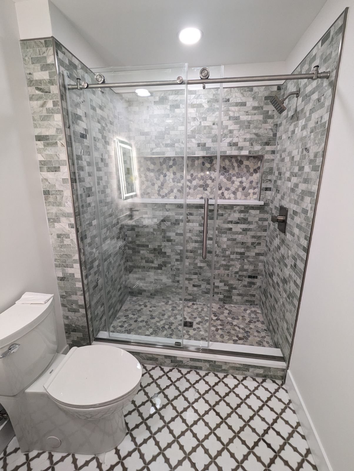 Modern bathroom with a glass-enclosed shower featuring gray stone tile walls, a built-in niche, and patterned tile floor.