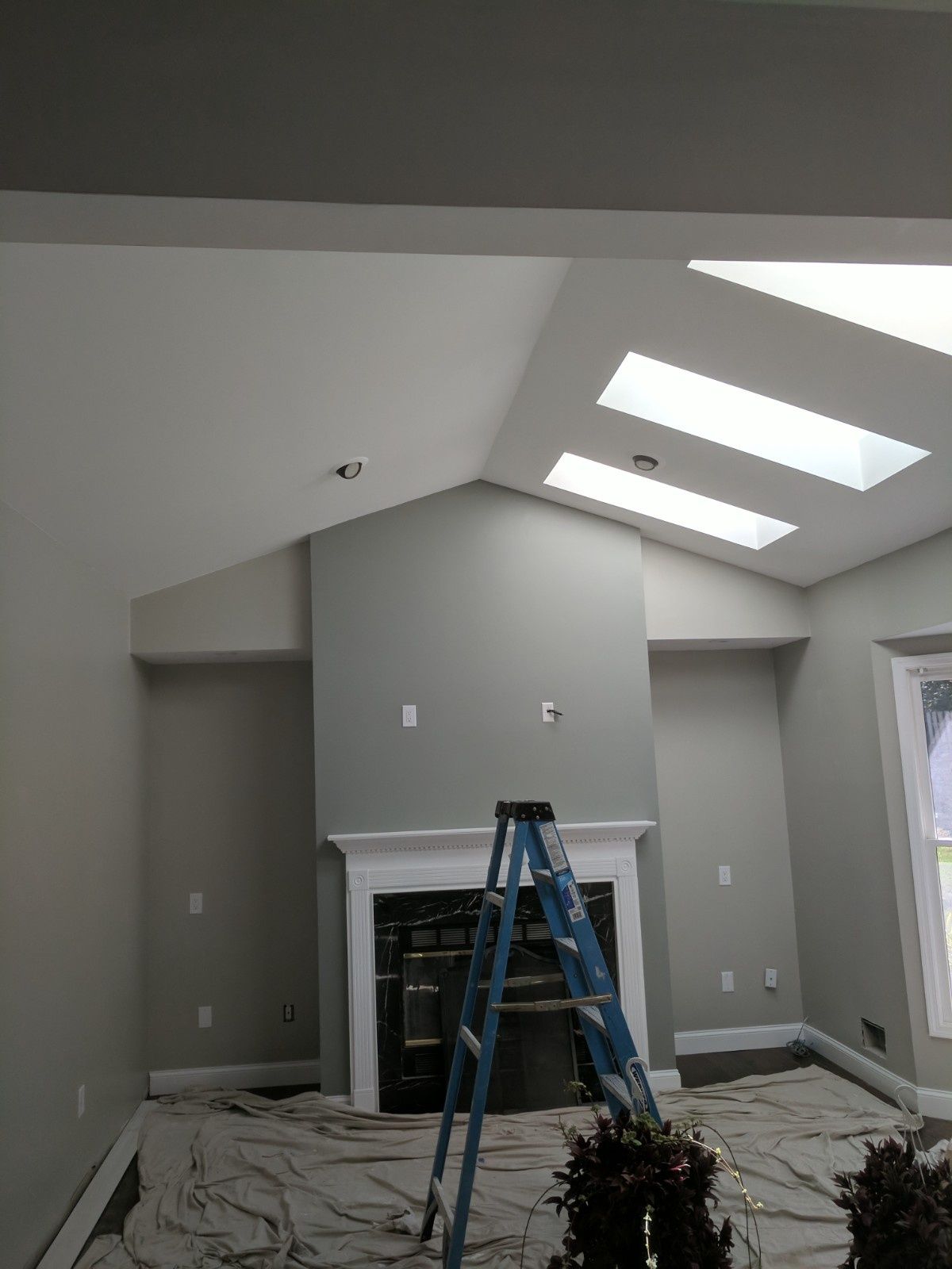 A blue step ladder stands on a drop cloth in front of a fireplace in a room with light gray walls and two skylights.