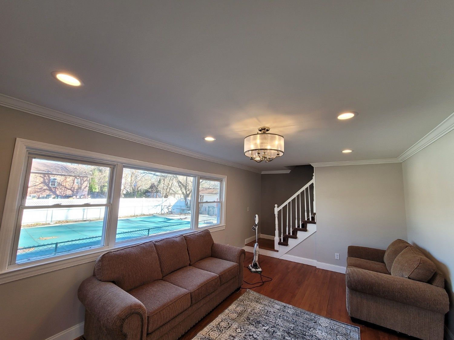 A living room with two brown sofas, wood flooring, a rug, a central chandelier, and a large window looking onto a pool.