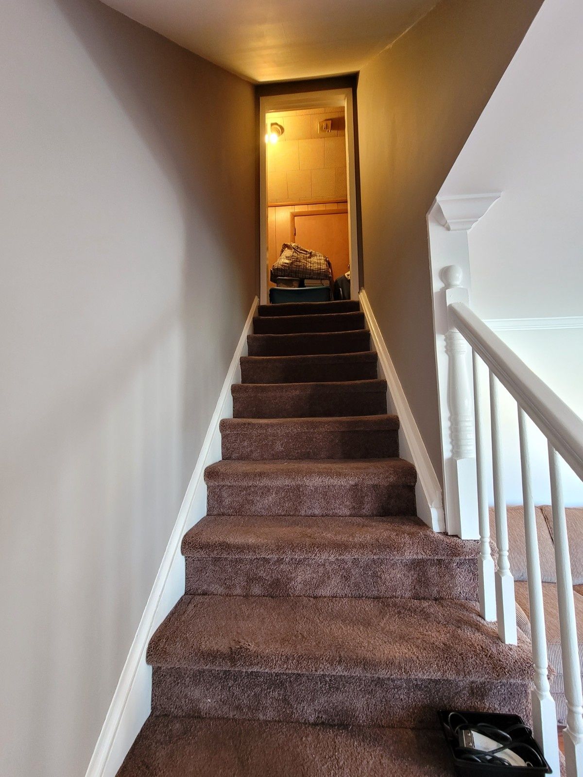 A narrow staircase covered in brown carpet leads upward toward a bright, open doorway at the top.