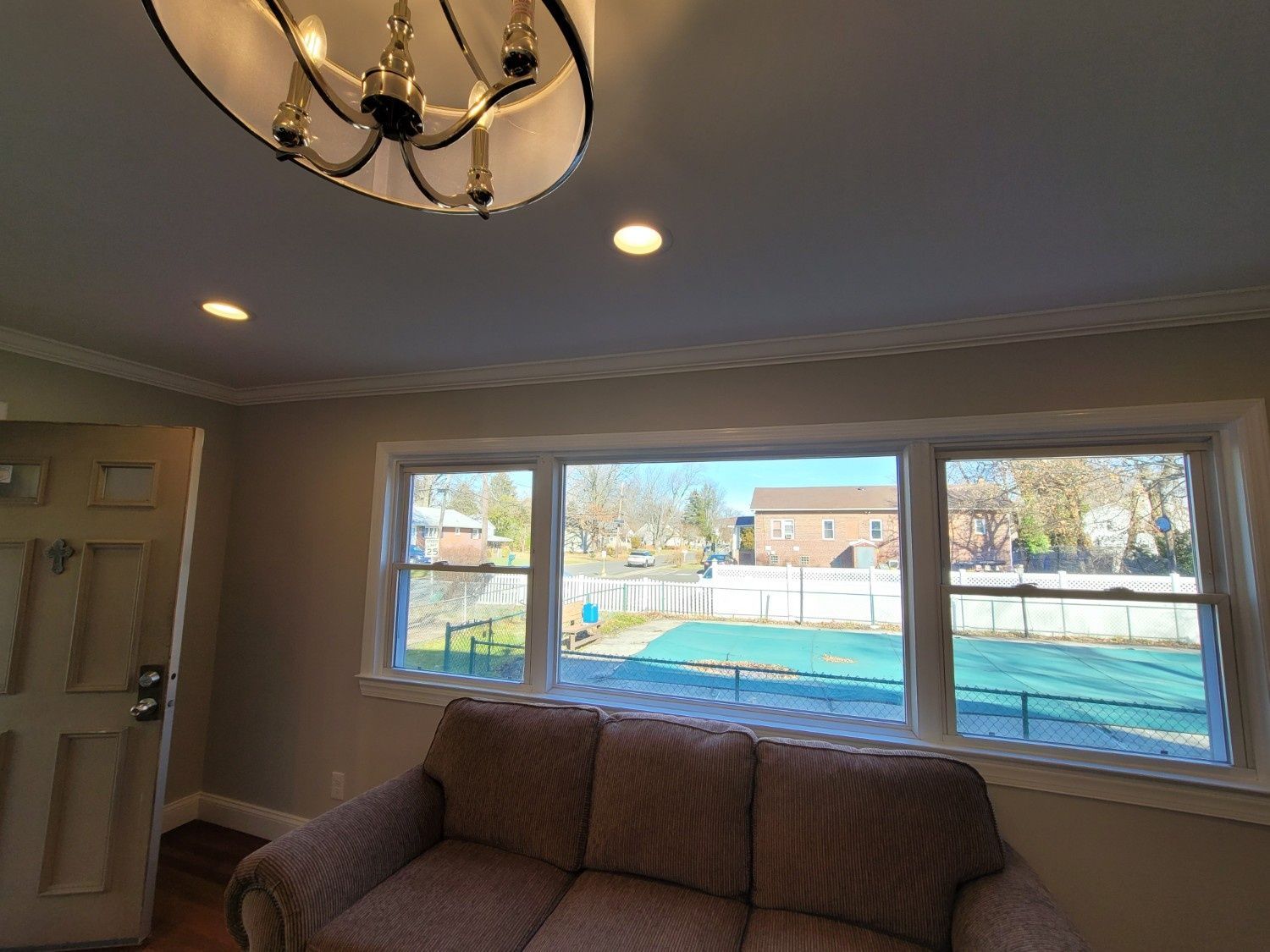 A living room with a beige sofa, a circular chandelier, recessed lighting, and a window view of a yard with a pool.