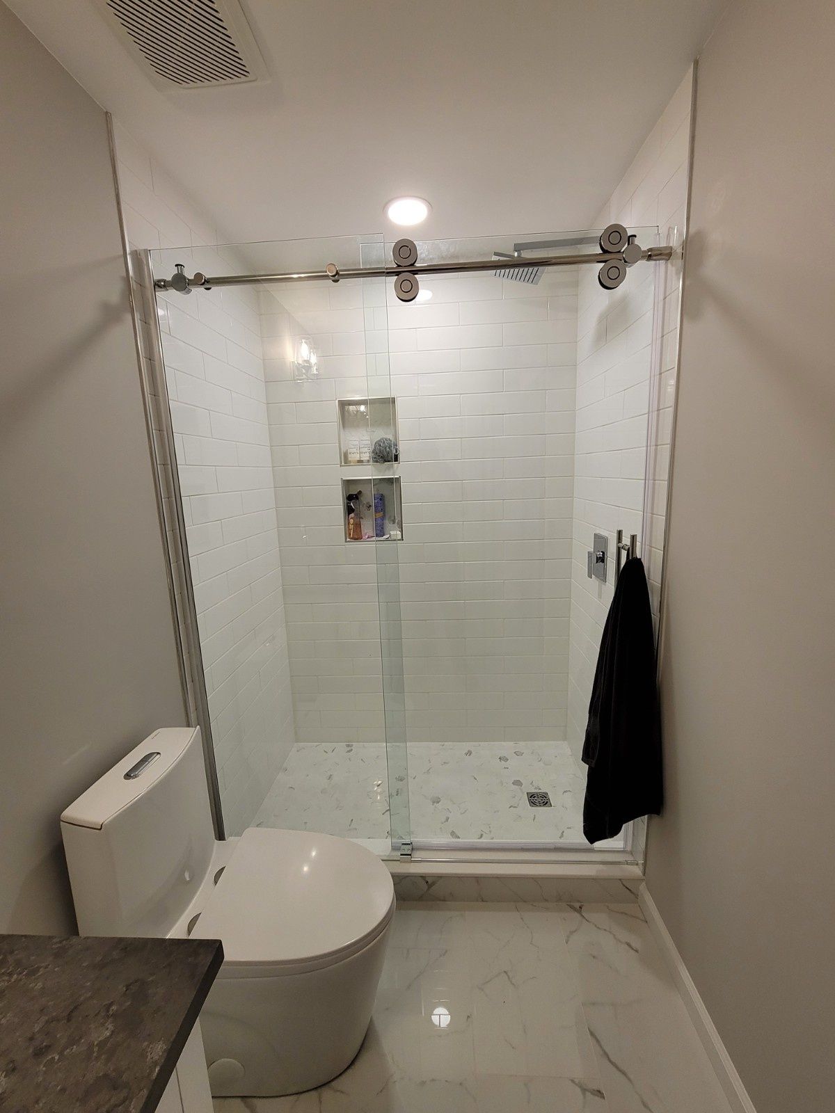 A modern bathroom featuring a white toilet next to a glass-enclosed shower with white tiled walls and marble floor tiles.