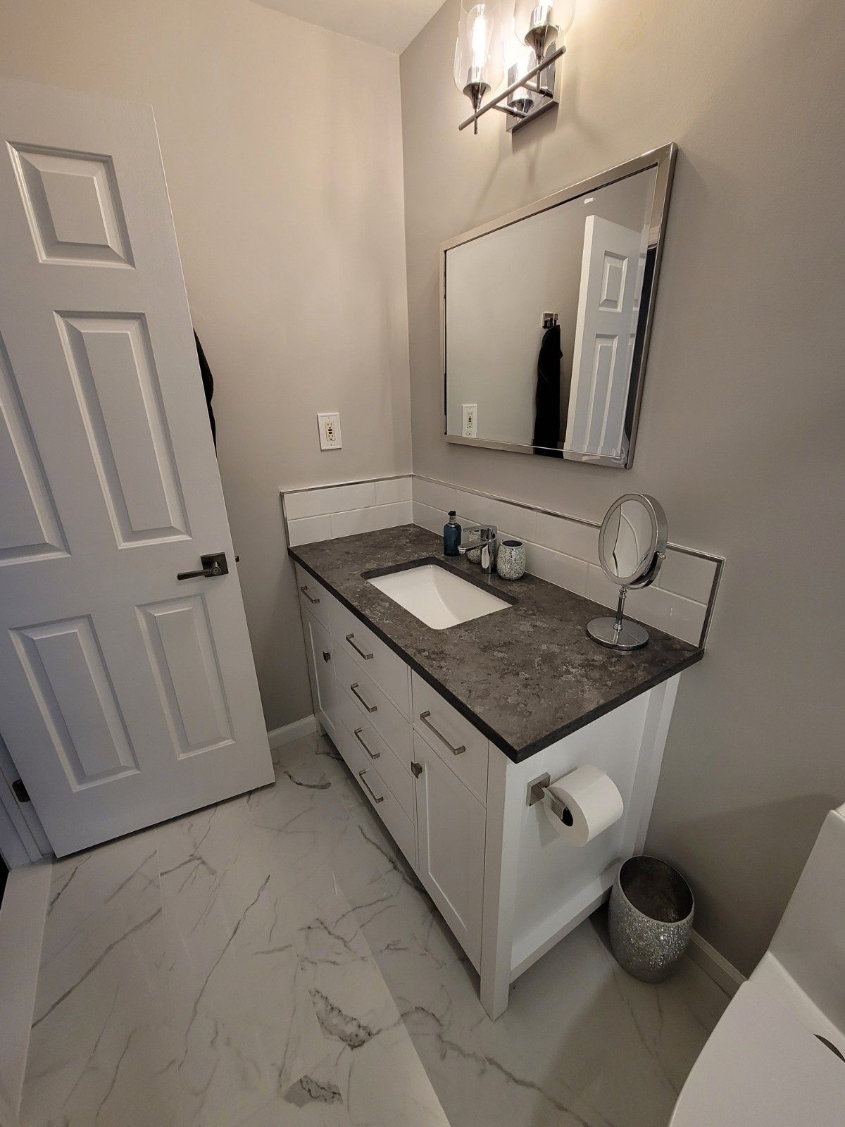 A bathroom with a white vanity, a dark gray countertop, a sink, a mirror, and white marble-patterned tile flooring.
