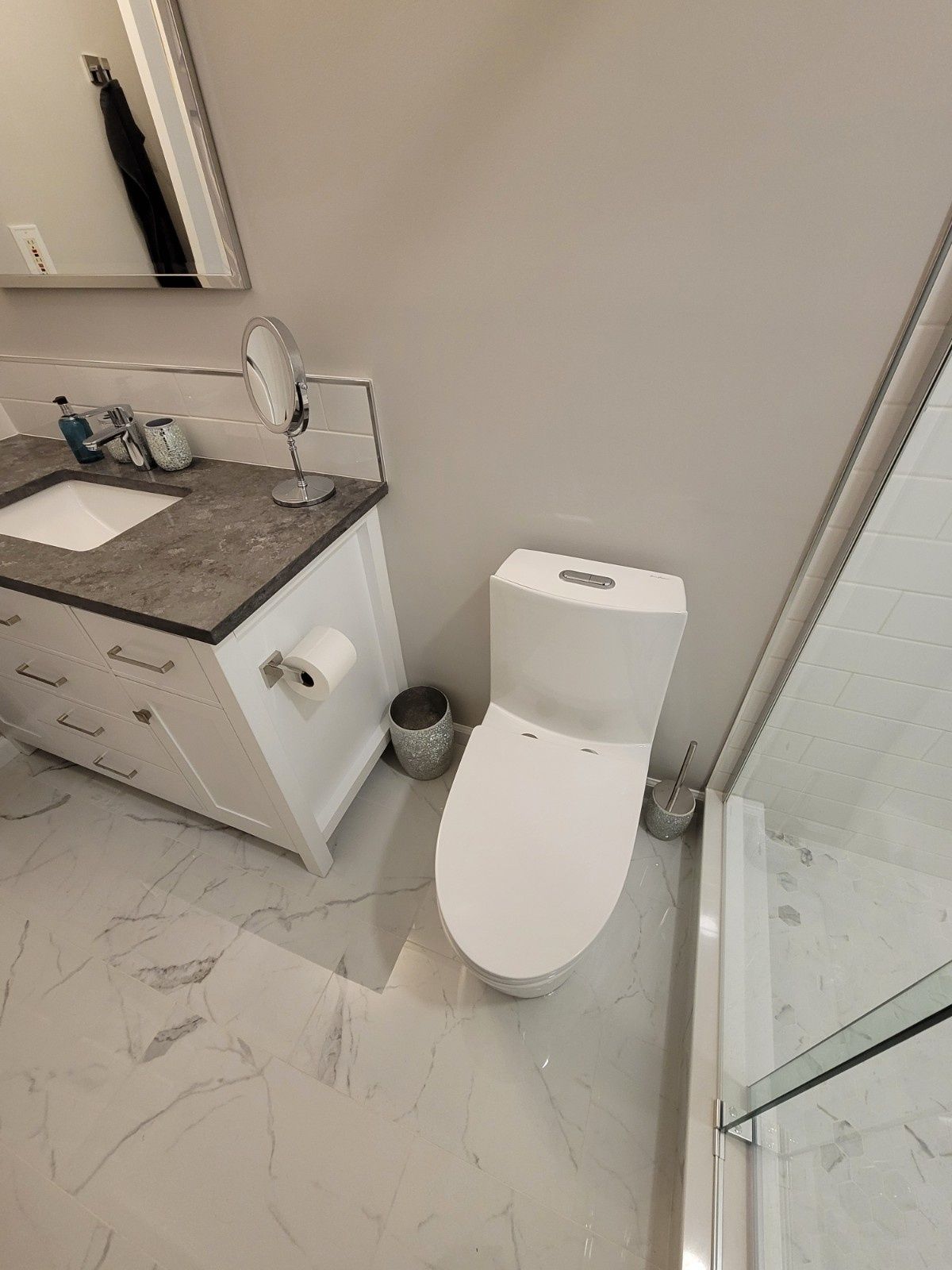 A high-angle view of a bathroom vanity with a gray stone top, next to a white toilet and a glass shower enclosure.