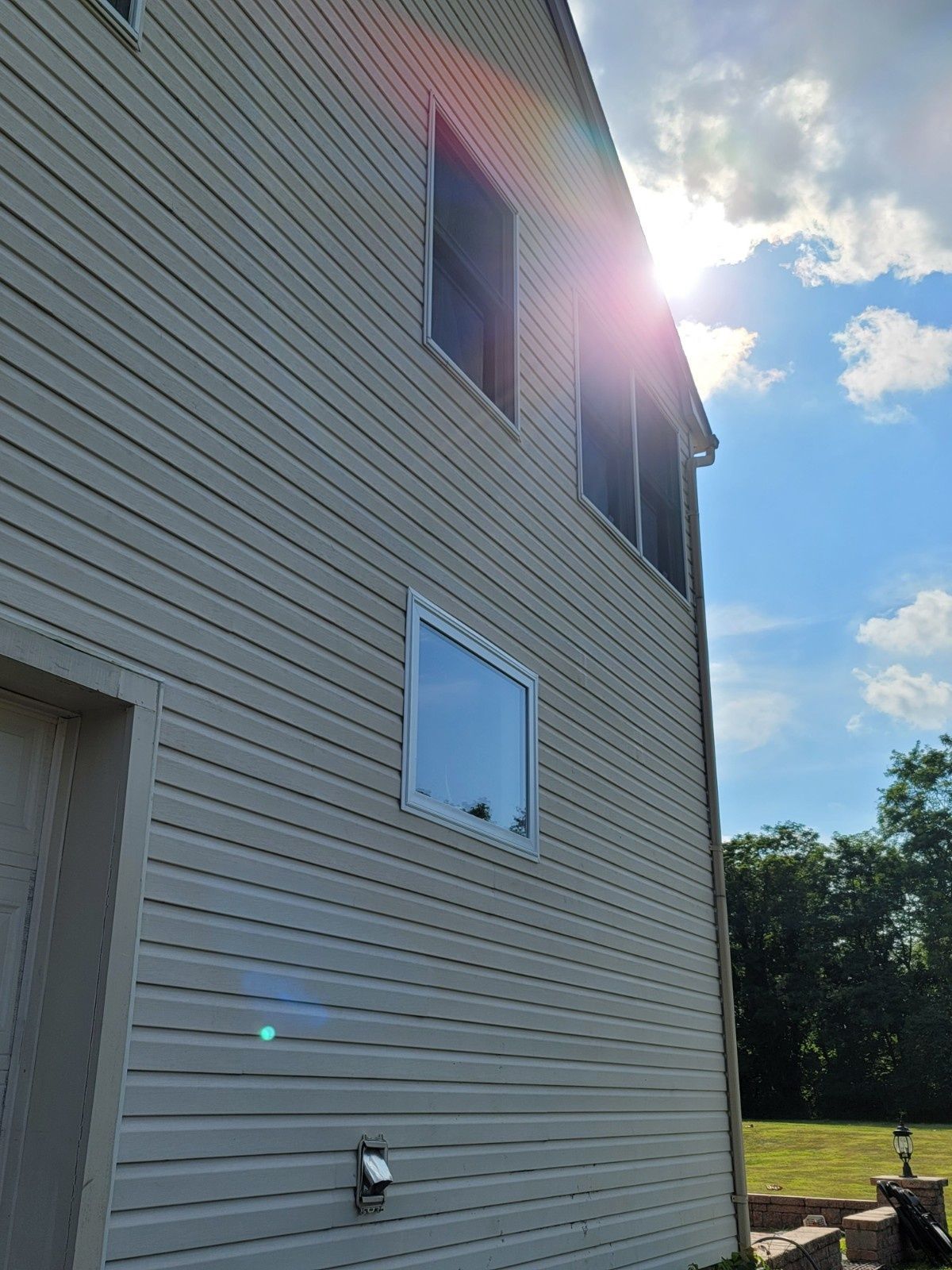 A low-angle view of a beige-sided house exterior with two windows and a bright sun flare in a blue sky.