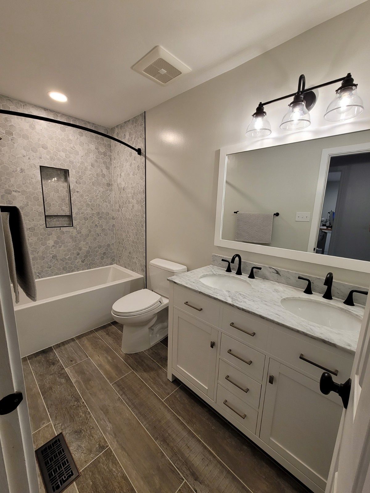 A modern bathroom with a white double-sink vanity, a tiled bathtub with a shower curtain, and wood-look tile flooring.