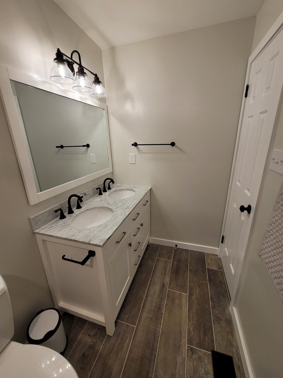 A bathroom vanity with a white cabinet, marble top, dual sinks, black fixtures, a large mirror, and wood-look tile floors.