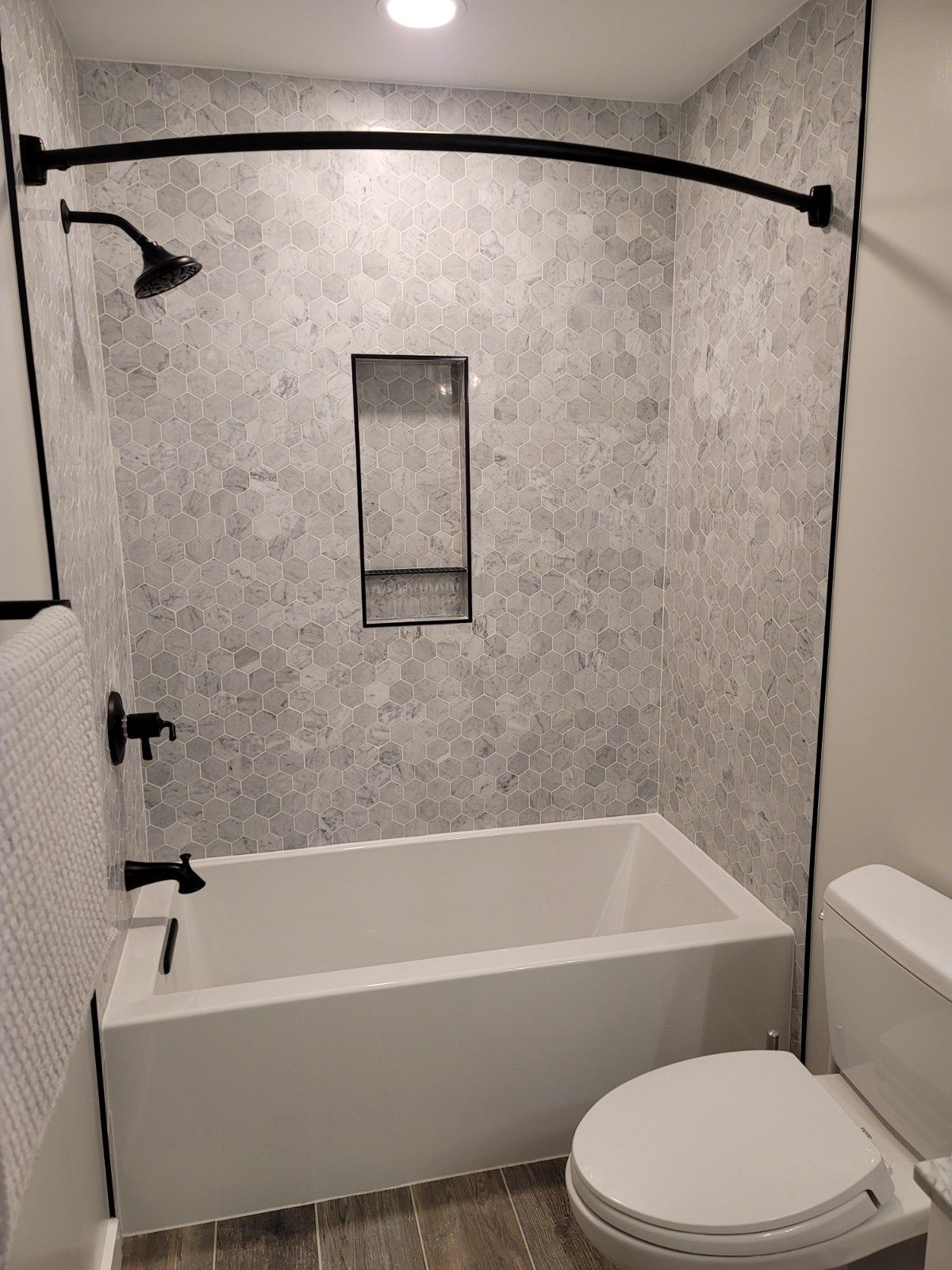 A bathroom featuring a white bathtub, a tiled shower wall with a recessed niche, matte black fixtures, and a curved rod.