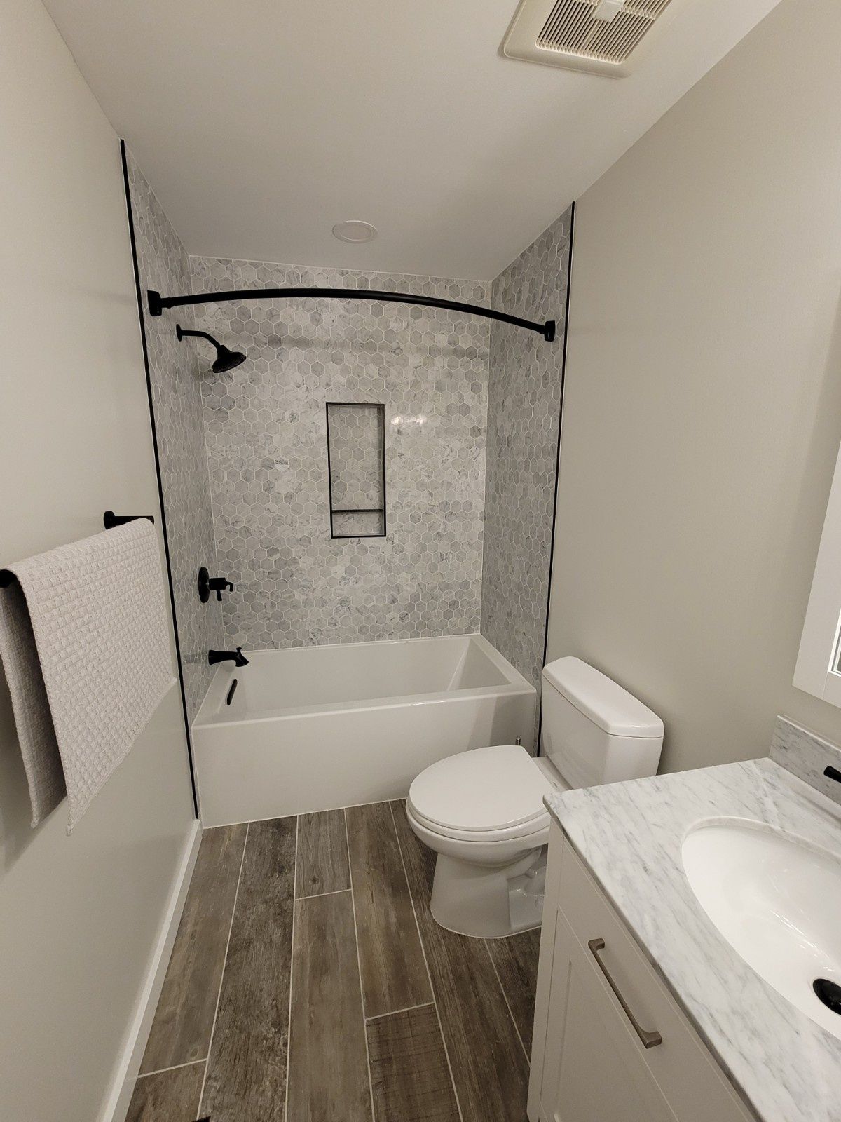 A modern bathroom featuring a white bathtub, gray marble-patterned tile, matte black fixtures, and wood-look floor tiles.