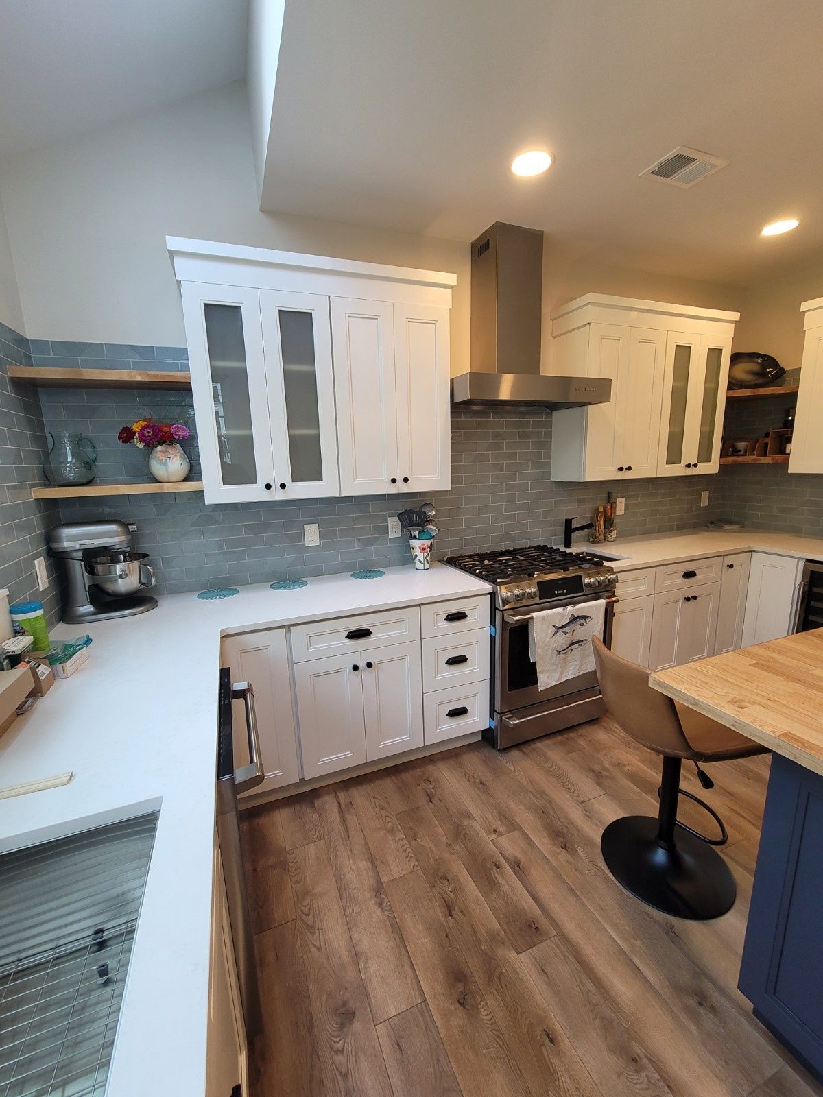 A bright kitchen with white cabinets, light countertops, wood-look flooring, stainless appliances, and open wood shelving.