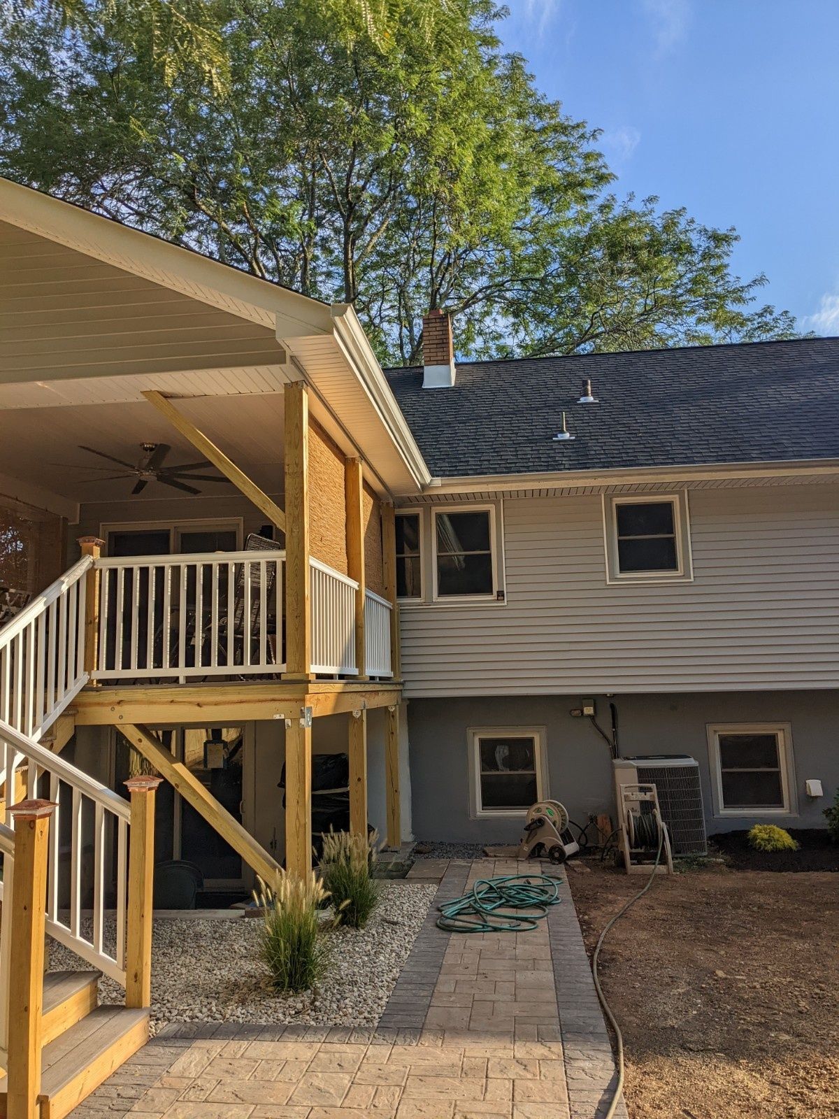 A multi-level backyard exterior showing a raised wood deck with white railings, a brick facade, and a stone walkway.