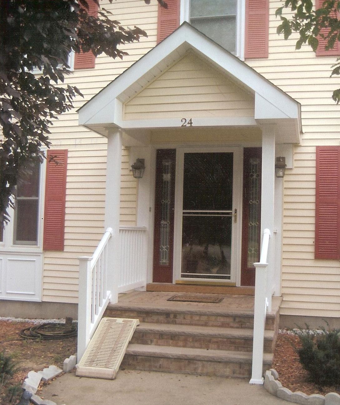 A front entryway of a yellow house with a gabled porch roof, red shutters, and a portable ramp on the stone stairs.