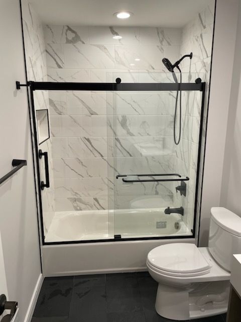 A modern bathroom featuring a white bathtub, black sliding glass doors, marble-patterned tile walls, and a white toilet.