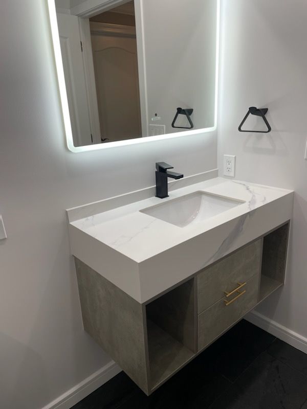 A wall-mounted bathroom vanity with a white stone top, black faucet, and gray cabinetry under a lit, frameless mirror.