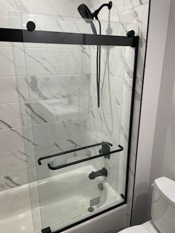 A glass shower door with a black frame and hardware installed over a white bathtub with white marble-patterned tile walls.