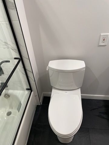 A white toilet positioned in a bathroom next to a glass shower enclosure with a black frame and a wall-mounted outlet.