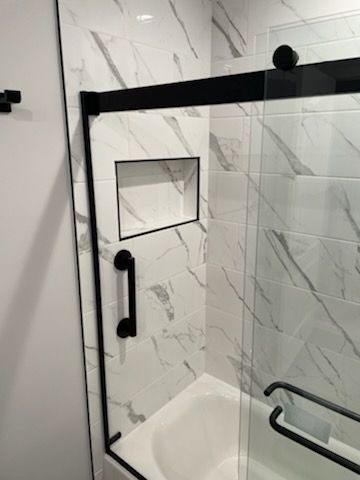 A bathroom shower featuring white marble-patterned tiles, a black-framed glass door, a black grab bar, and a wall-recessed niche.