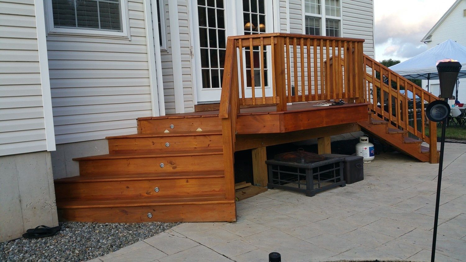 A wooden deck with a staircase leads to a house entrance above a concrete patio, with a fire pit stored underneath.