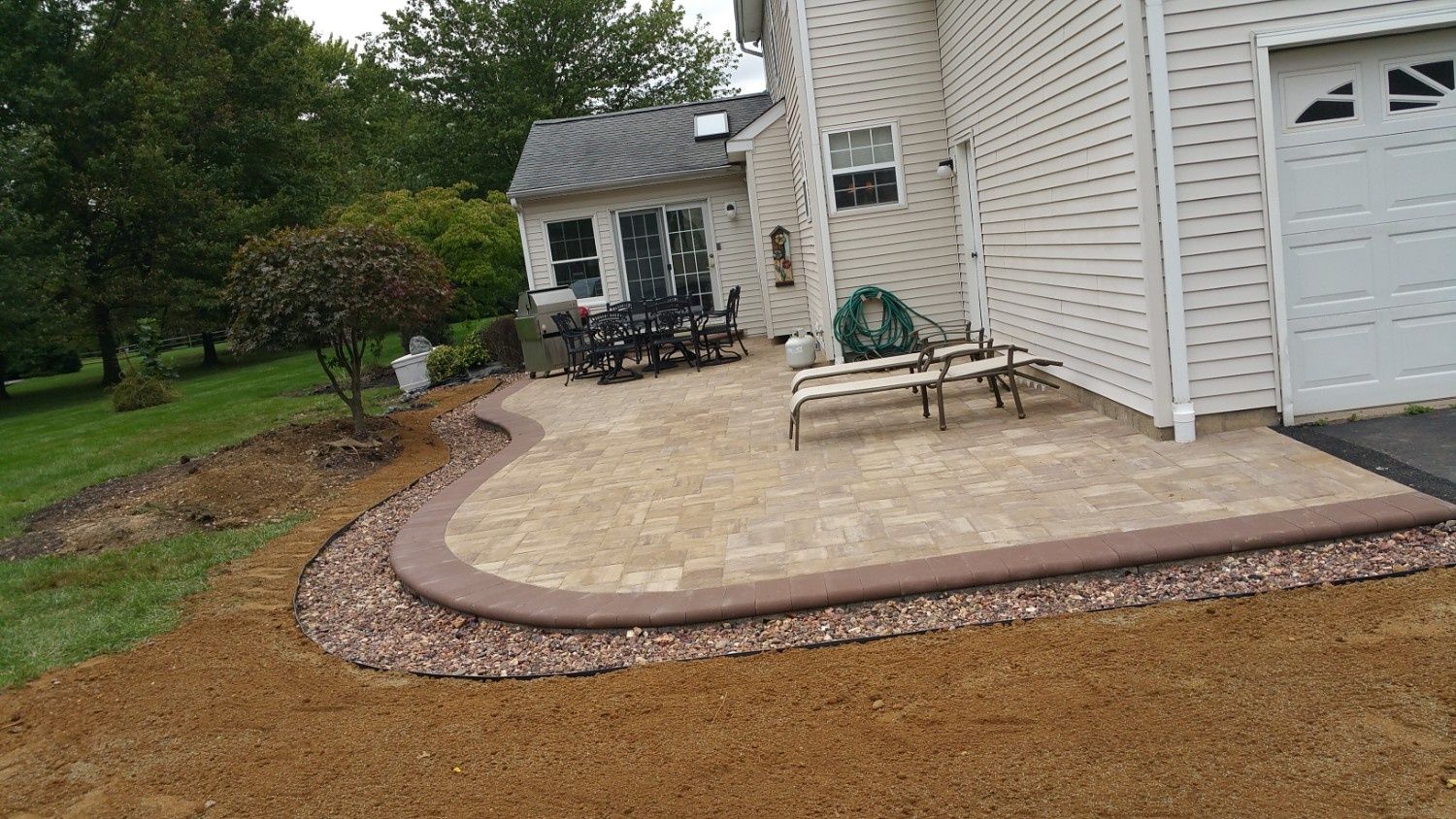 A tan stone patio with a curved brown border, surrounded by gravel and bare soil next to a beige house with a garage.