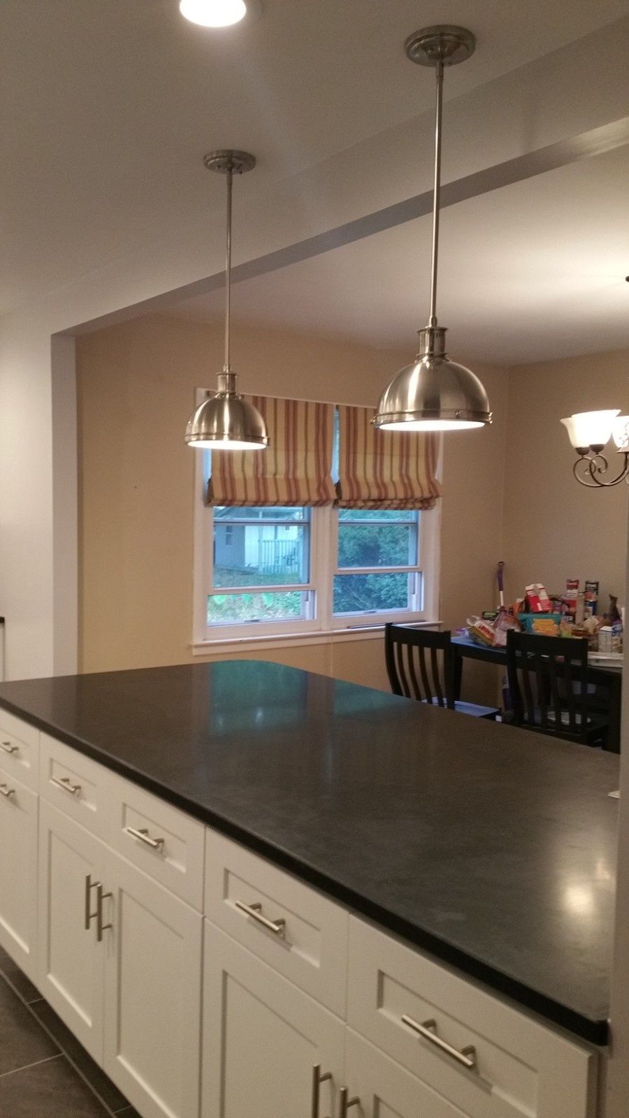 A kitchen island with a dark countertop and white cabinets, lit by two pendant lights, with a dining area in the background.