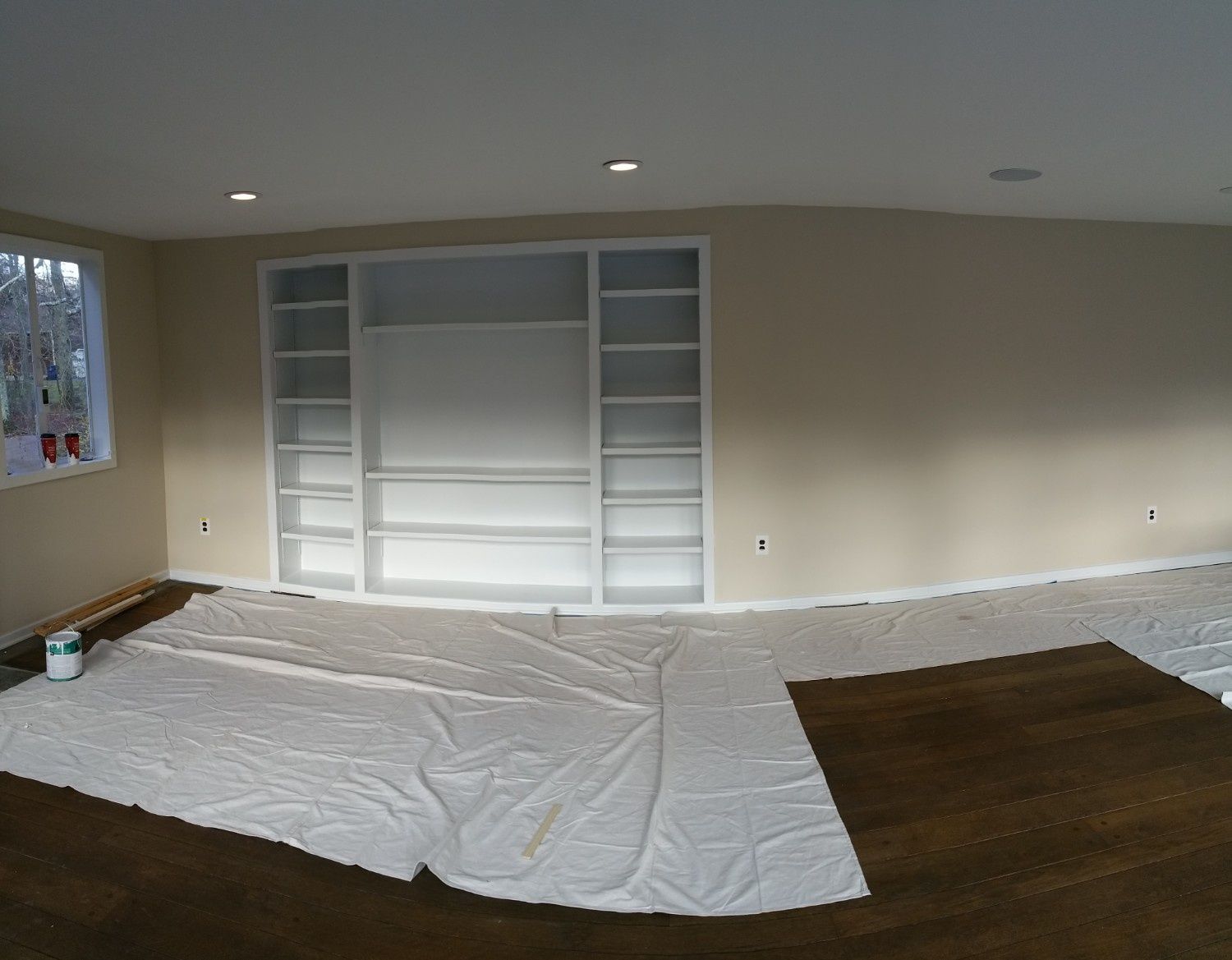 A panoramic view of an unfinished room with tan walls, white built-in shelving, and dark hardwood floors under a drop cloth.
