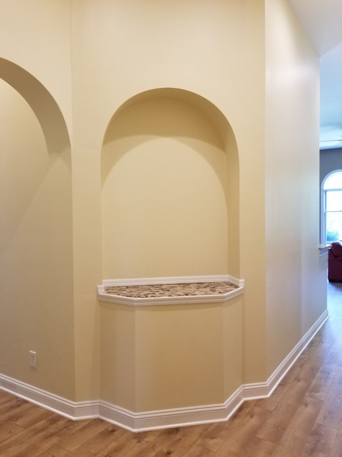 An arched wall niche featuring a small, tiled ledge, set against beige walls in a brightly lit hallway with wood flooring.