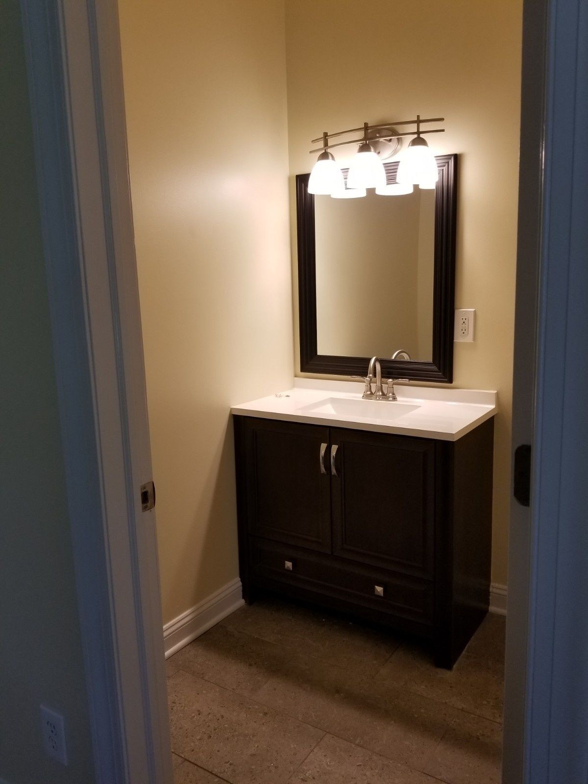 A small bathroom featuring a dark wood vanity with a white countertop, sink, mirror, and four-bulb light fixture.