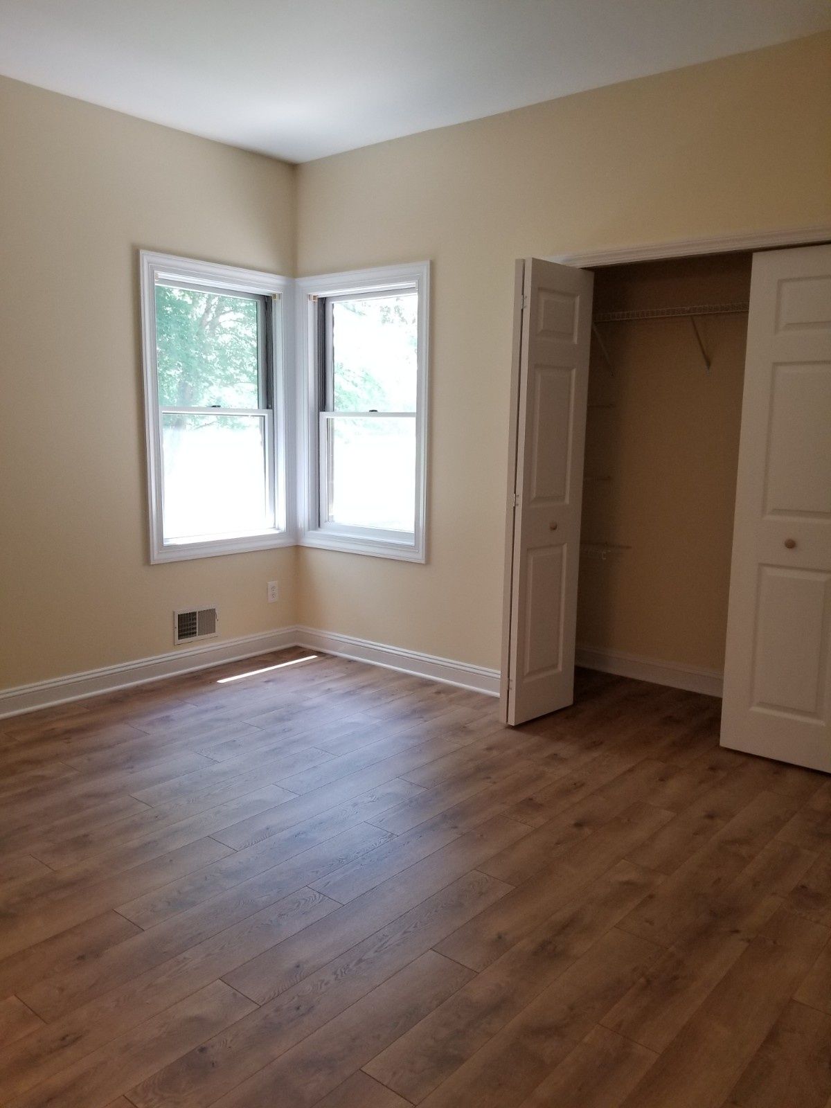 An empty bedroom with light beige walls, wood-look flooring, two windows in the corner, and an open bi-fold closet door.