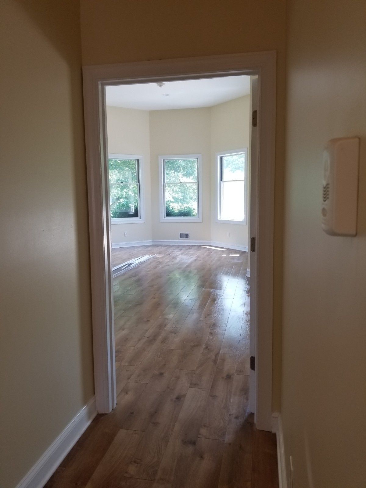 A hallway view through a doorway into a room with wood flooring, light-colored walls, and a bay window.