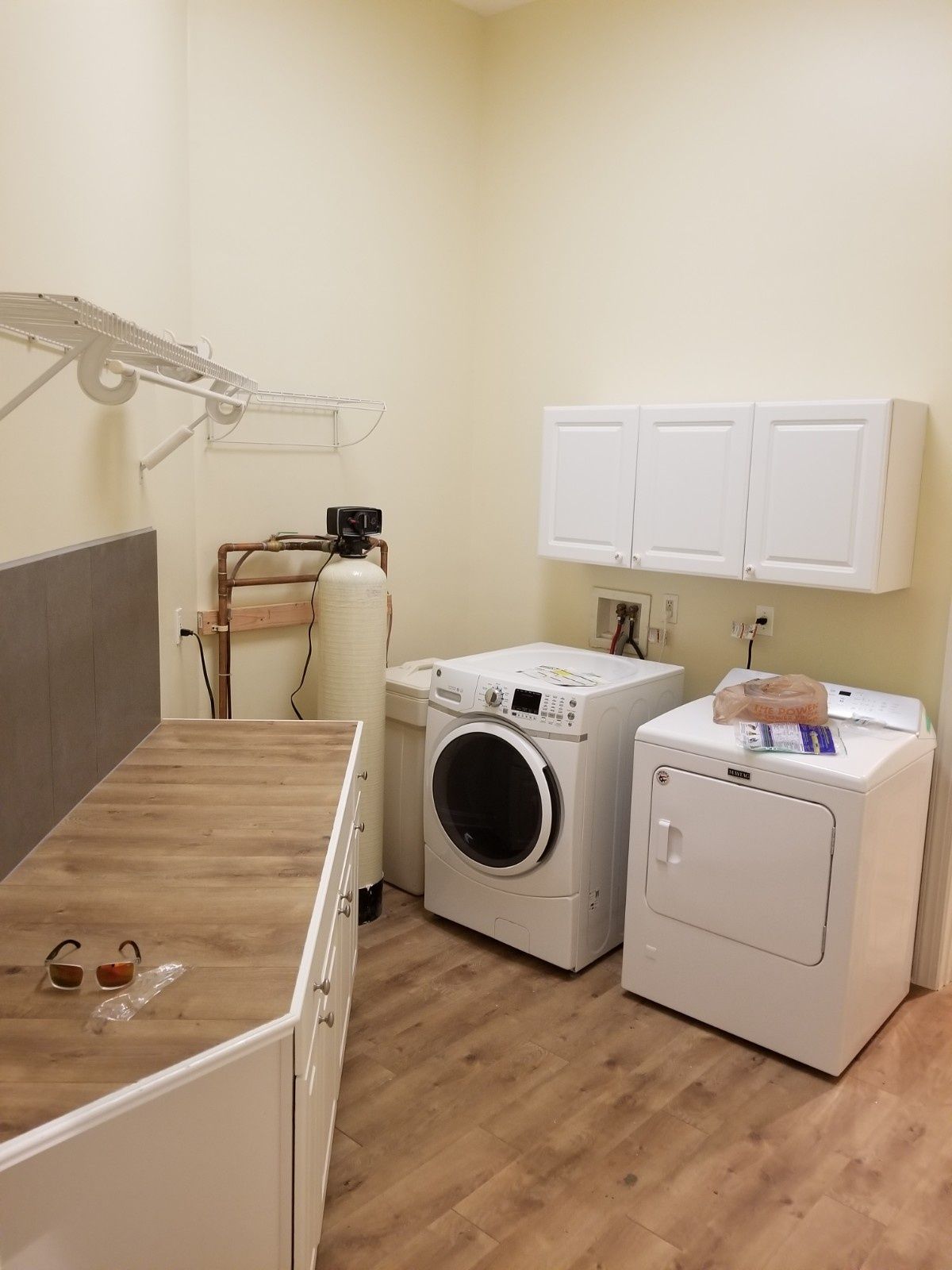 A laundry room with a washer, dryer, wooden countertop, overhead cabinets, and a white water filtration tank.