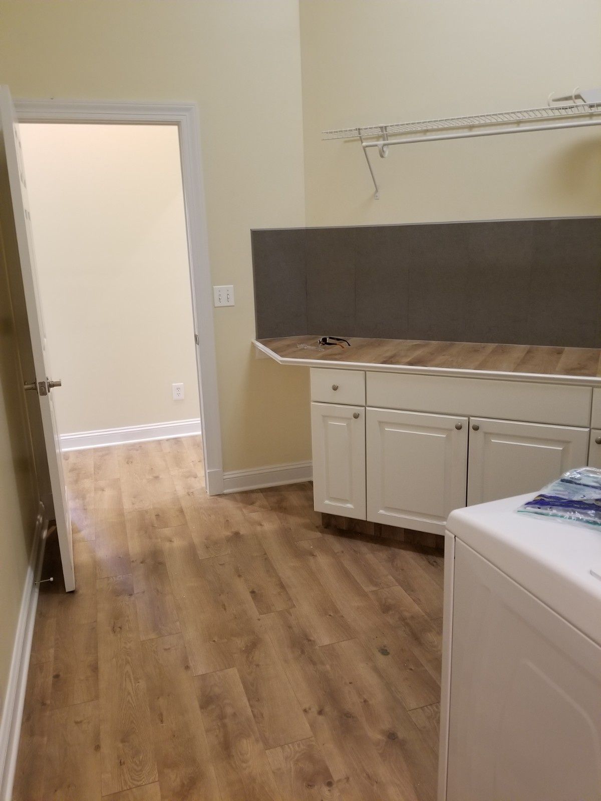 Laundry room with light wood-look flooring, white cabinets, a brown backsplash, and an open doorway to another room.
