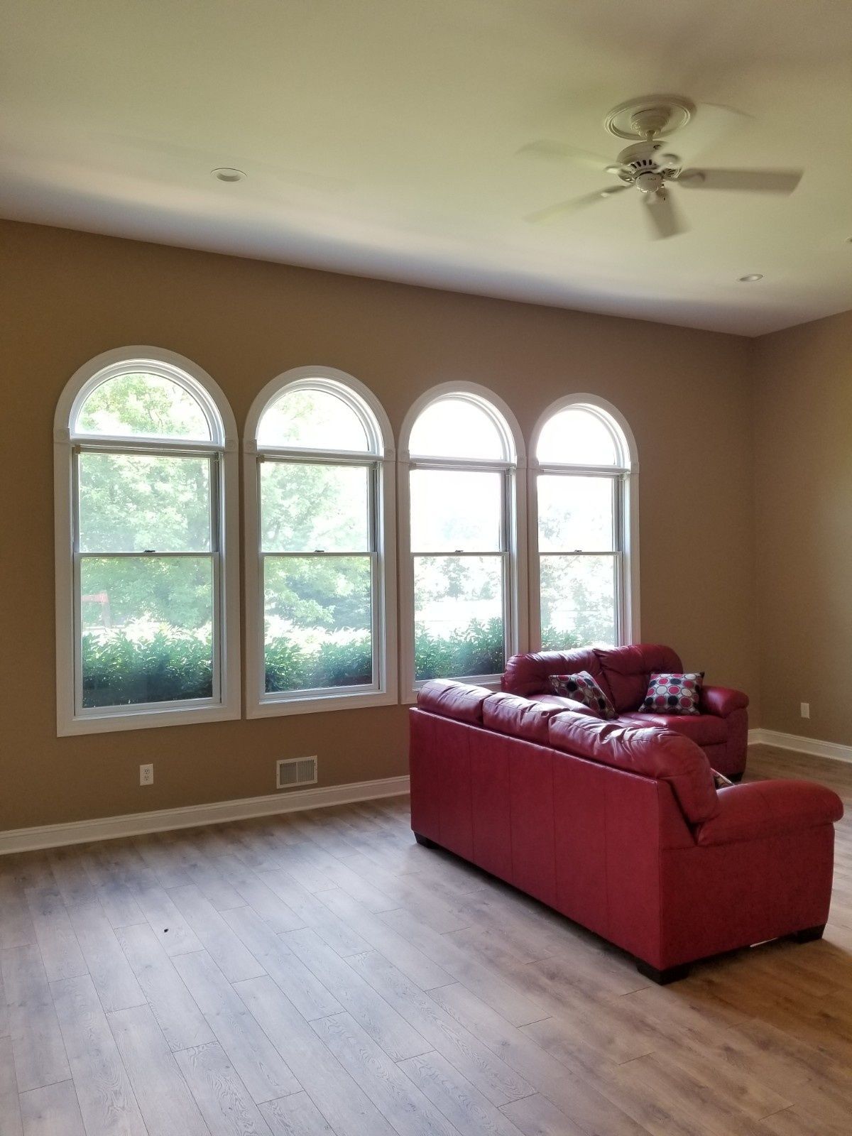 A red leather sofa sits in an empty living room with light wood floors, beige walls, and four arched windows.